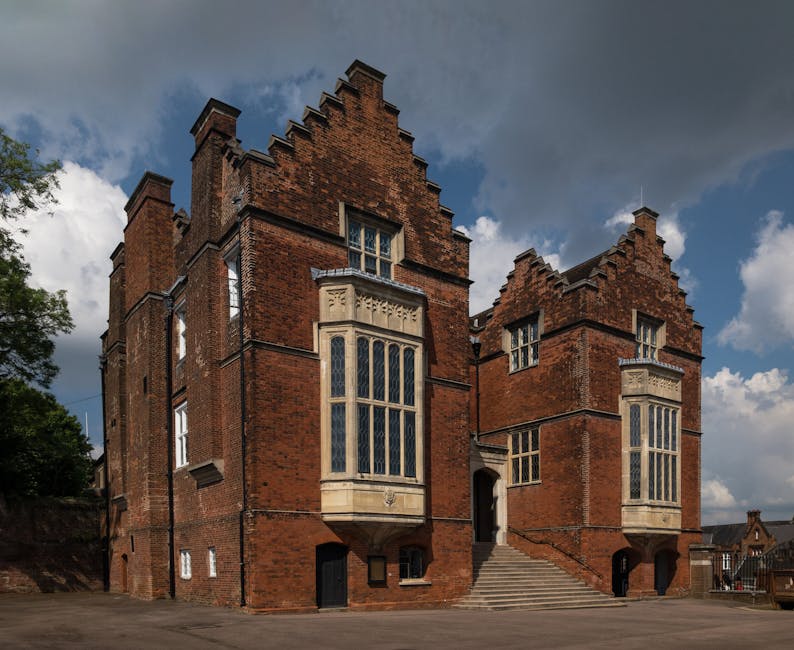 Photograph of a historic red brick building with a distinctive Gothic architectural style, featuring large, tall windows with stone frames, some with decorative mullions. The building has stepped gables and staircase leading up to an arched entrance, with a clear blue sky and white clouds overhead. The surrounding area includes a paved courtyard and some greenery. The scene reflects well-maintained exteriors, showcasing cleanliness and preservation, which aligns with the professional cleaning services offered by Cleaners Harrow for surface maintenance and deep cleaning, as referenced on the page titled 'Carpet cleaning near Harrow School HA1 trusted teams, Harrow'.
