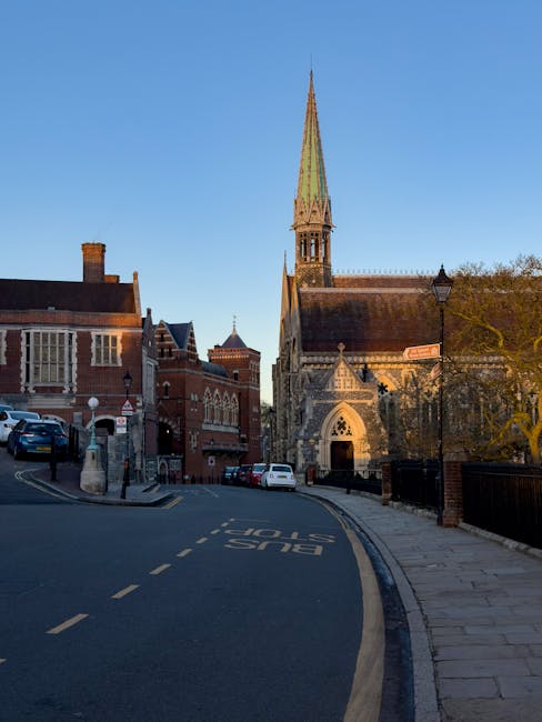 Photograph of a historic red brick building with a distinctive Gothic architectural style, featuring large, tall windows with stone frames, some with decorative mullions. The building has stepped gables and staircase leading up to an arched entrance, with a clear blue sky and white clouds overhead. The surrounding area includes a paved courtyard and some greenery. The scene reflects well-maintained exteriors, showcasing cleanliness and preservation, which aligns with the professional cleaning services offered by Cleaners Harrow for surface maintenance and deep cleaning, as referenced on the page titled 'Carpet cleaning near Harrow School HA1 trusted teams, Harrow'.