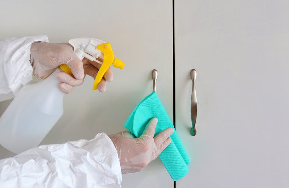 A person wearing white gloves and a protective suit is using a yellow spray bottle to apply cleaning solution to a light blue cloth. They are cleaning a grey kitchen cabinet door with a curved metal handle. The cabinet surface is smooth and clean, reflecting good hygiene. The background shows similar grey cabinets with sleek, metal handles, and soft, natural lighting illuminates the area. This image exemplifies surface cleaning and sanitisation as part of domestic cleaning services provided by Cleaners Harrow, focusing on maintaining a hygienic and spotless kitchen environment, aligned with tips for affordable cleaning prices in Wealdstone HA3.