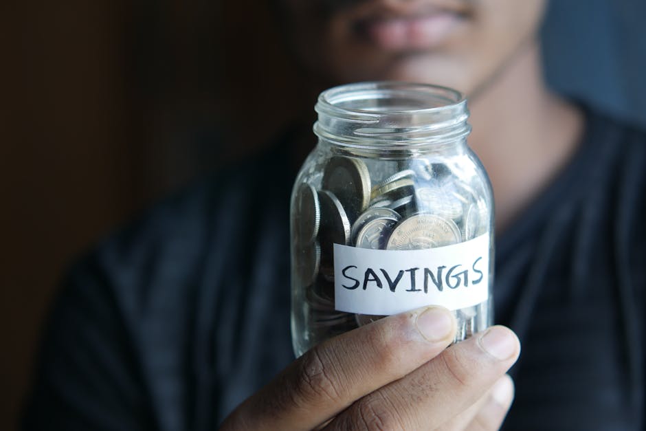 A person holding a clear glass jar filled with various coins, with a white paper label that reads 'SAVINGS' attached to the front of the jar. The background is blurred, focusing on the hand and jar, indicating a concept of financial savings or budgeting related to domestic cleaning expenses on the website cleaners-harrow.com, specifically for the 'Domestic cleaning prices Wealdstone HA3' page by Cleaners Harrow. The scene emphasizes the idea of saving money through expert cleaning services, with soft lighting highlighting the jar and coins.