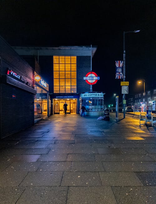 The image shows the entrance to Pinner station at night, with a well-lit glass facade and illuminated signage, including the London Underground roundel and a digital display board. The pavement is wet, reflecting the lights from streetlamps and surrounding buildings. To the left, storefronts with closed shutters are visible, while on the right, a street lamp and British flags hang from a pole. Several pedestrians are walking towards the station entrance, and the overall scene emphasizes the cleanliness and maintained appearance of the station exterior, consistent with professional cleaning and maintenance standards. This setting aligns with the services provided by Cleaners Harrow for property hygiene and surface cleaning in the Pinner station area.