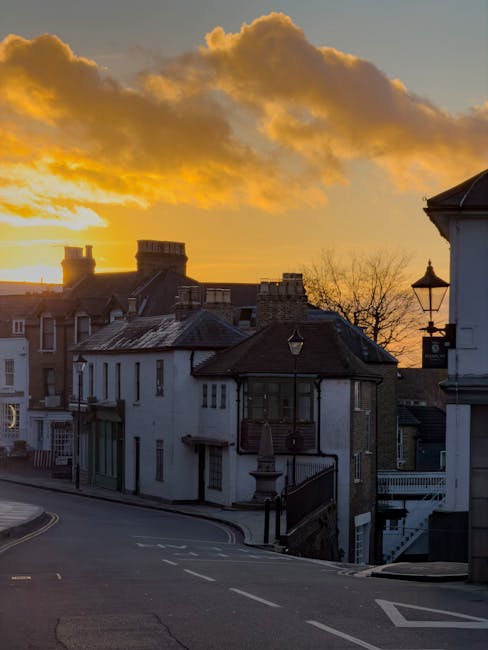 A view of residential buildings in Pinner station area Harrow during sunset, featuring a mix of brick and stucco exteriors, pitched tiled roofs, and chimneys. The street is quiet with paved roads and black metal fencing along the property boundaries. Warm orange and yellow hues illuminate the sky with scattered clouds, creating a serene atmosphere. The scene is well-lit by natural evening light, highlighting the clean and well-maintained appearance of the neighborhood, consistent with the high standards of domestic surface cleaning and maintenance promoted by Cleaners Harrow for end of tenancy cleaning services in the Harrow area.
