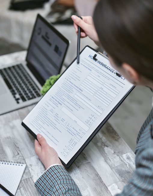 A person reviewing a printed insurance form with a pen, seated at a wooden desk alongside a laptop displaying a webpage. The desk surface appears clean and organized, with natural lighting illuminatin