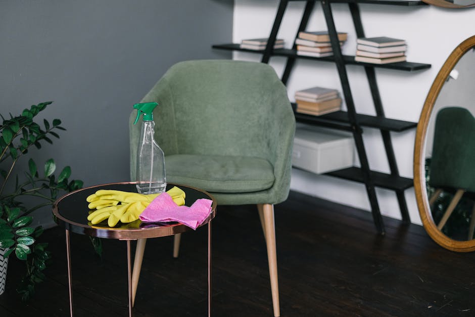 A green upholstered armchair is situated in a room with a dark wooden floor and a white wall. In front of the armchair, there is a round black side table with a copper-colored trim, holding a pair of yellow rubber cleaning gloves, a pink cleaning cloth, and a transparent spray bottle with a green nozzle. To the left of the armchair, there is a potted green plant with broad leaves. Behind the chair, a black ladder-style bookshelf with several neatly stacked books is visible, and a white storage box is placed on one of the shelves. To the right of the room, a large oval mirror leans against the wall, reflecting part of the space. The overall scene suggests a maintained, clean, and organized domestic environment with natural lighting emphasizing the tidy appearance, appropriate for the surface cleaning and deep cleaning topics associated with Cleaners Harrow and their focus on sanitisation and hygiene as referenced in the page about hidden cleaning charges in Harrow.