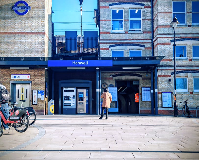 The image shows the entrance to Hanwell station, a historic brick building with a modern black canopy above the ticket machines, situated in Harrow. The façade features a mix of beige and red bricks, with rectangular white-framed windows on the upper floors. A woman wearing a beige coat and jeans stands in front of the ticket machines, possibly purchasing tickets. To the left, there are rental bikes branded with Uber and VCS parked near a designated bike rack. The station's signage, including the blue 'Elizabeth Line' sign, is prominently displayed above the entrance. The platform area is clean and well-maintained, illuminated by a classic street lamp on the right, under a bright, clear sky. Cleaners Harrow specializes in surface cleaning, deep cleaning, and sanitisation for commercial and residential spaces, ensuring hygiene and cleanliness in transportation hubs like this.