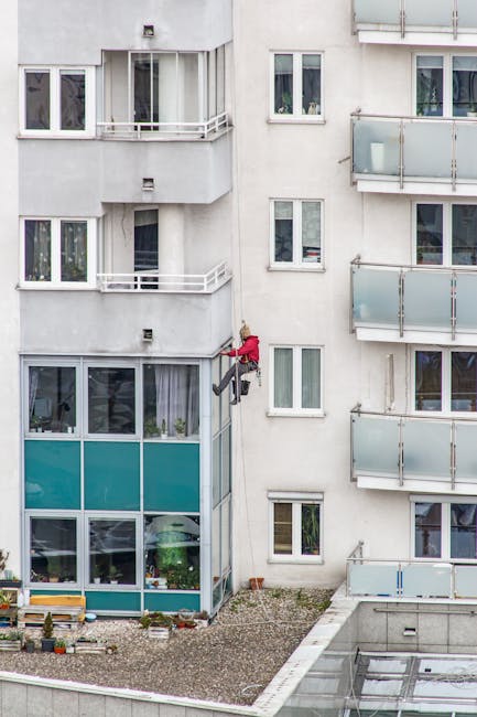 Two professional window cleaners from Cleaners Harrow are suspended on ropes outside a modern multi-storey building, performing surface cleaning and sanitisation of the glass windows. They are equippe
