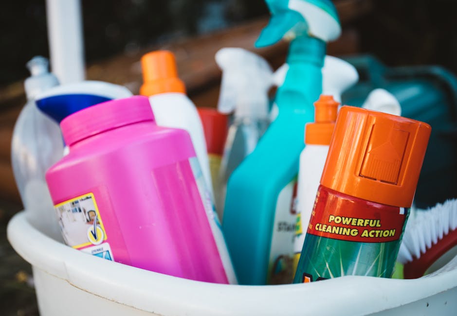 A row of domestic cleaning bottles of various sizes and colors, including blue, orange, yellow, blue, and pink, are arranged on a white surface against a plain, light gray wall. The bottles are made o