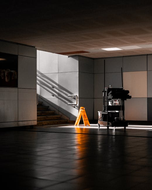 An indoor commercial space featuring a tiled floor with a matte finish, illuminated by natural light streaming in from an unseen source. In the background, a staircase with stainless steel handrails ascends along a wall composed of smooth, light-colored panels. Adjacent to the staircase, a cleaning cart equipped with various cleaning supplies and tools is positioned, alongside a bright yellow wet floor caution sign standing on the tiled surface. The overall scene highlights a clean, well-maintained environment suitable for office or business centre cleaning, with surfaces appearing spotless and surfaces well-organized, emphasizing the importance of hygiene and surface cleanliness for Hatch End business centres as promoted by Cleaners Harrow.