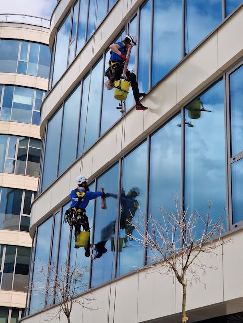 Two professional window cleaners from Cleaners Harrow are performing surface cleaning on the exterior glass facade of a modern multi-story office building in Hatch End, Harrow. The workers are secured with safety harnesses and ropes while working on separate floors. One cleaner, dressed in a blue uniform and helmet, is wiping the large glass windows with a cloth, ensuring a streak-free shine, while the other, in a red uniform and helmet, is using a spray bottle to apply cleaning solution to the glass surface. The building features reflective, tinted glass panels framed by light-colored concrete walls, with clear blue sky and scattered cloud reflections visible in the windows. The scene is set during daytime with natural sunlight illuminating the surfaces. The image reflects professional exterior cleaning processes by the crew from Cleaners Harrow, emphasizing the importance of building maintenance and hygiene through such detailed window care.