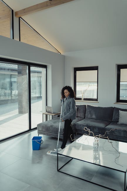 A woman with curly hair cleaning a modern living room using a mop, with a blue bucket on the light gray tiled floor. The room features large floor-to-ceiling glass sliding doors with black frames, providing natural light and views of an outdoor patio. The interior includes a dark gray fabric sectional sofa, a glass coffee table with decorative branches, and three smaller windows with black frames on the white walls. The ceiling is sloped with exposed wooden beams, and the space appears clean and well-maintained, reflecting the domestic cleaning services offered by Cleaners Harrow, emphasizing surface cleaning and sanitisation for a hygienic environment.